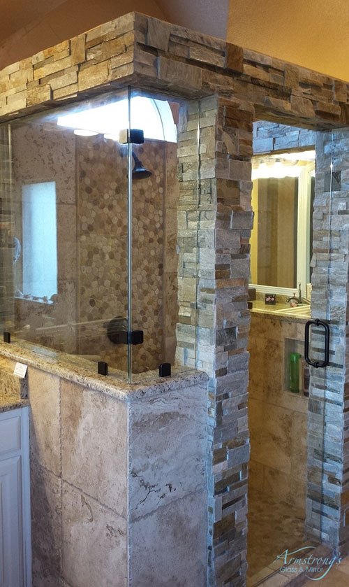 A modern bathroom with a stone and glass walk-in shower, featuring beige tiles and a rainfall showerhead. A vanity with a mirror and lighting is visible through the shower's glass wall.