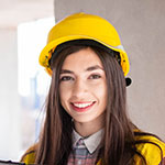 A young woman wearing a yellow construction helmet and a plaid shirt smiles at the camera. She has long brown hair and stands in a partially constructed building.