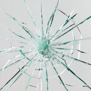 Close-up of shattered glass with cracks radiating outward from a central impact point against a light gray background. The broken pieces create sharp, jagged lines in various directions.